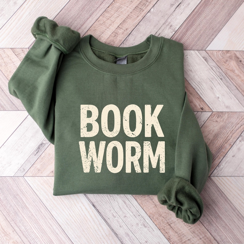 May include: An olive green sweatshirt with the words "BOOK WORM" printed in a distressed, off-white font. The sweatshirt is folded, with the sleeves cuffed. The background is a light wood grain pattern.