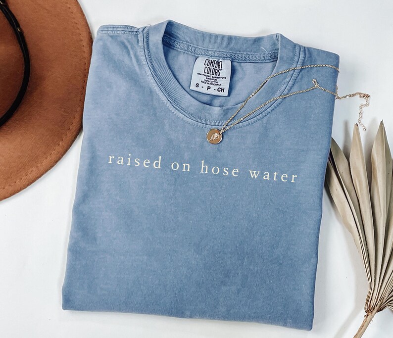 May include: A blue t-shirt with the phrase "raised on hose water" in white lettering. A gold necklace with a pendant rests on the shirt. The shirt is folded and placed on a white surface, with a brown hat and dried plant in the background.