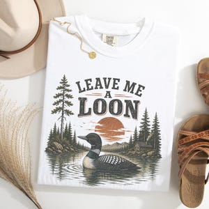 Pu&ograve; includere: Una t-shirt bianca con una stampa grafica di un colimbo che nuota su un lago, con una foresta, montagne e un sole al tramonto sullo sfondo. Il testo "LEAVE ME A LOON" &egrave; stampato sopra l'illustrazione in un carattere serif scuro e usurato.