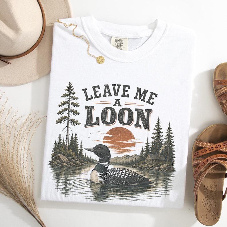 Pu&ograve; includere: Una t-shirt bianca con una stampa grafica di un colimbo che nuota su un lago, con una foresta, montagne e un sole al tramonto sullo sfondo. Il testo "LEAVE ME A LOON" &egrave; stampato sopra l'illustrazione in un carattere serif scuro e usurato.