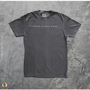 May include: A charcoal gray t-shirt with the phrase "raised on hose water" printed in white across the chest. The shirt has a crew neck and short sleeves. The fabric appears soft and slightly faded.