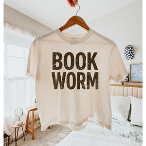 May include: Cream-colored t-shirt with the words "BOOK WORM" printed in a distressed, dark brown font. The shirt is hanging on a wooden hanger.