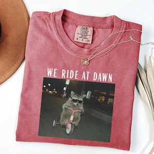 Puede incluir: Camiseta roja Comfort Colors con la frase "WE RIDE AT DAWN" impresa en blanco. La camiseta presenta un gr&aacute;fico de un mapache con gafas de sol montando un triciclo rosa. Un collar plateado con un colgante est&aacute; sobre la camiseta.