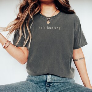 May include: Dark gray t-shirt with the text "he's hunting" in a simple, white font. The shirt has a slightly faded appearance and a classic crew neck. The model is wearing blue jeans and a gold necklace.