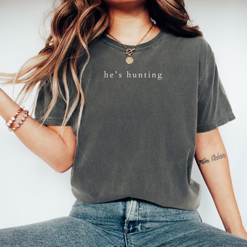 May include: Dark gray t-shirt with the text "he's hunting" in a simple, white font. The shirt has a slightly faded appearance and a classic crew neck. The model is wearing blue jeans and a gold necklace.