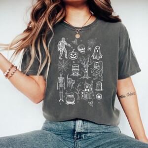 May include: Dark grey t-shirt with a white Halloween-themed design. The illustrations include a mummy, pumpkin, skull, spiderweb, ghost, Frankenstein's monster, werewolf, skeleton, cat, haunted house, and cauldron.