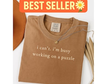 May include: A brown Comfort Colors t-shirt with the text "i can't. i'm busy working on a puzzle." A silver necklace with a small pendant rests on the shirt. The words "BEST SELLER" are in an orange banner.