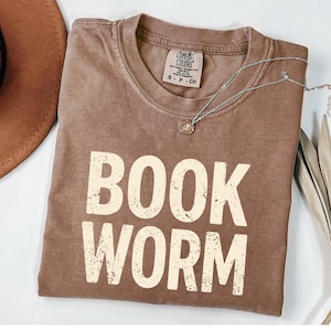May include: A brown t-shirt with the words "BOOK WORM" printed in a distressed, off-white font. The shirt has a crew neck and short sleeves. A silver necklace with a small pendant is draped across the shirt. The shirt is folded on a white surface.