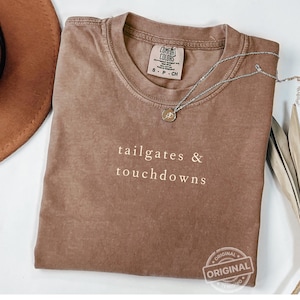 Może przedstawiać: Brązowa koszulka Comfort Colors z białym napisem "tailgates & touchdowns". Srebrny naszyjnik z wisiorkiem leży na koszulce. Koszulka jest złożona i ma metkę przy dekolcie. Częściowo widoczny brązowy kapelusz.
