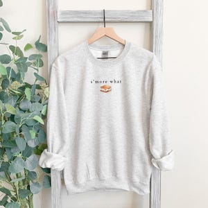 May include: A light gray sweatshirt with the text "s'more what" above a graphic of a s'more. The sweatshirt has a crew neck and long sleeves. The sleeves are cuffed at the wrists.