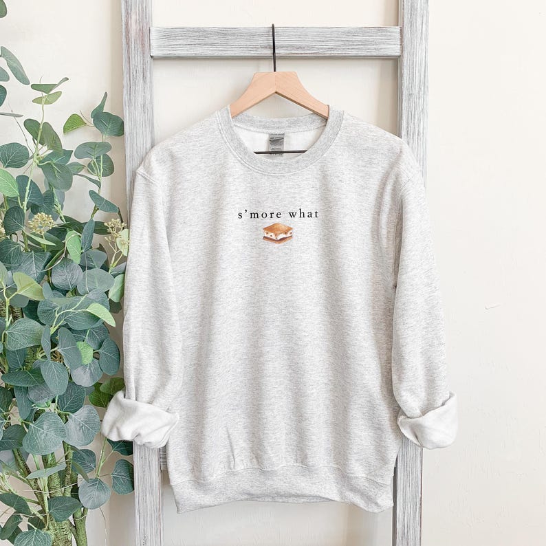 May include: A light gray sweatshirt with the text "s'more what" above a graphic of a s'more. The sweatshirt has a crew neck and long sleeves. The sleeves are cuffed at the wrists.