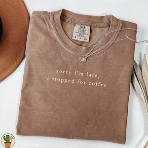 Puede incluir: Una camiseta marr&oacute;n Comfort Colors con el texto "sorry i'm late, i stopped for coffee" en blanco. Un collar plateado con un colgante circular descansa sobre la camiseta. La camiseta es talla S-P-CH.