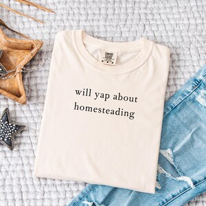 K&ouml;nnte beinhalten: Cremefarbenes T-Shirt mit dem Text "will yap about homesteading" in Schwarz. Das Shirt ist gefaltet und liegt auf einer strukturierten Oberfl&auml;che neben einer hellblauen Jeans und Sternornamenten.