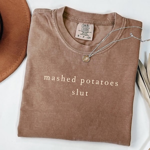 May include: A brown Comfort Colors t-shirt with the words "mashed potatoes slut" printed in a light brown color. A silver necklace with a small pendant is draped across the shirt. The shirt is folded neatly.