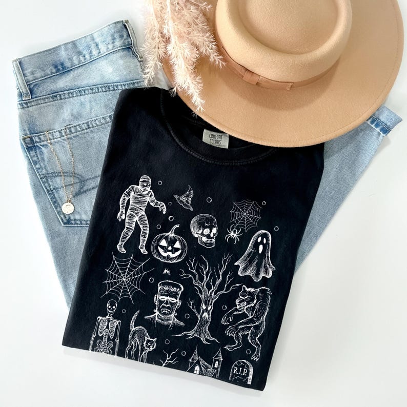 May include: A black t-shirt featuring a white Halloween design, alongside light-wash denim jeans, a beige hat, and a gold necklace. The graphic includes a mummy, pumpkin, skull, spiderweb, ghost, and other spooky elements.