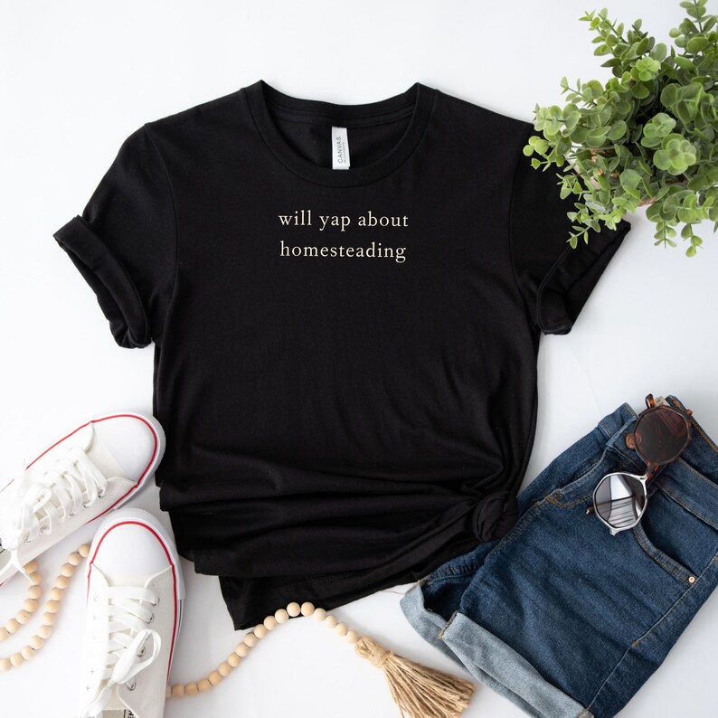 K&ouml;nnte beinhalten: Schwarzes T-Shirt mit dem Text "will yap about homesteading" in einfacher, wei&szlig;er Schrift. Das Shirt ist mit blauen Jeansshorts, wei&szlig;en Turnschuhen, Sonnenbrille und dekorativen Perlen versehen. Eine kleine gr&uuml;ne Pflanze ist ebenfalls zu sehen.
