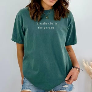 May include: A teal green t-shirt with the text "i'd rather be in the garden" in white. The shirt is a short-sleeved, casual style. The model is wearing denim shorts and jewelry.