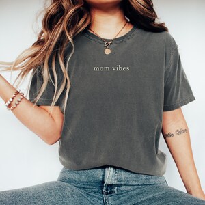May include: A woman wearing a dark gray t-shirt with the text "mom vibes" printed on the front. She is also wearing blue jeans and a gold necklace.