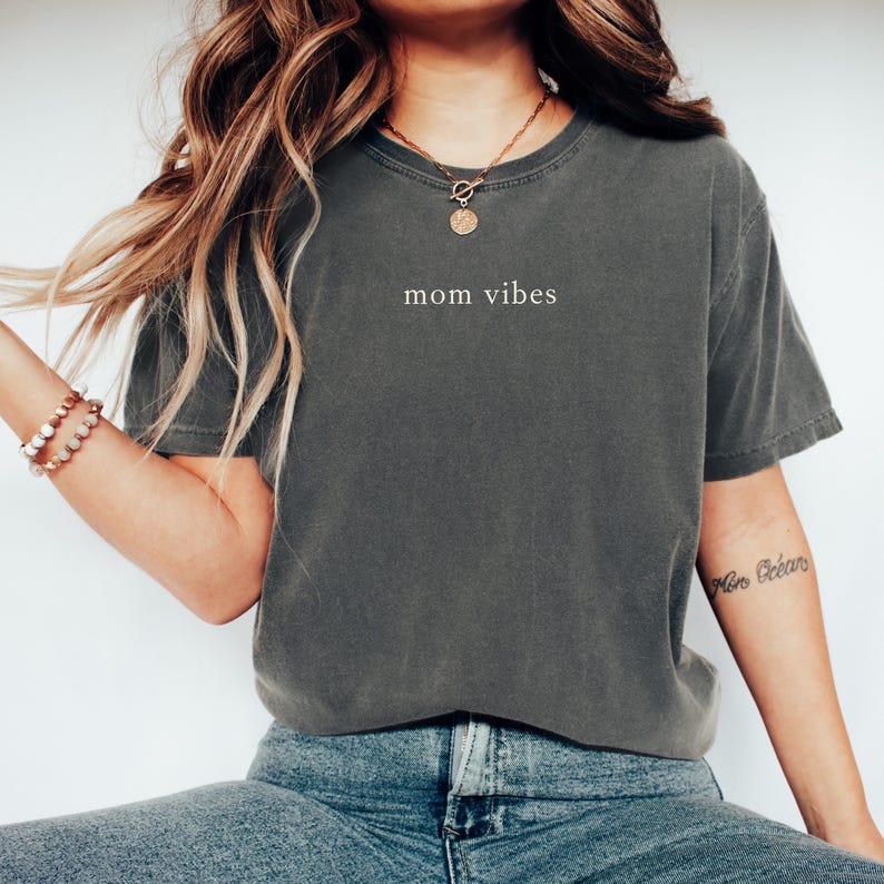 May include: A woman wearing a dark gray t-shirt with the text "mom vibes" printed on the front. She is also wearing blue jeans and a gold necklace.