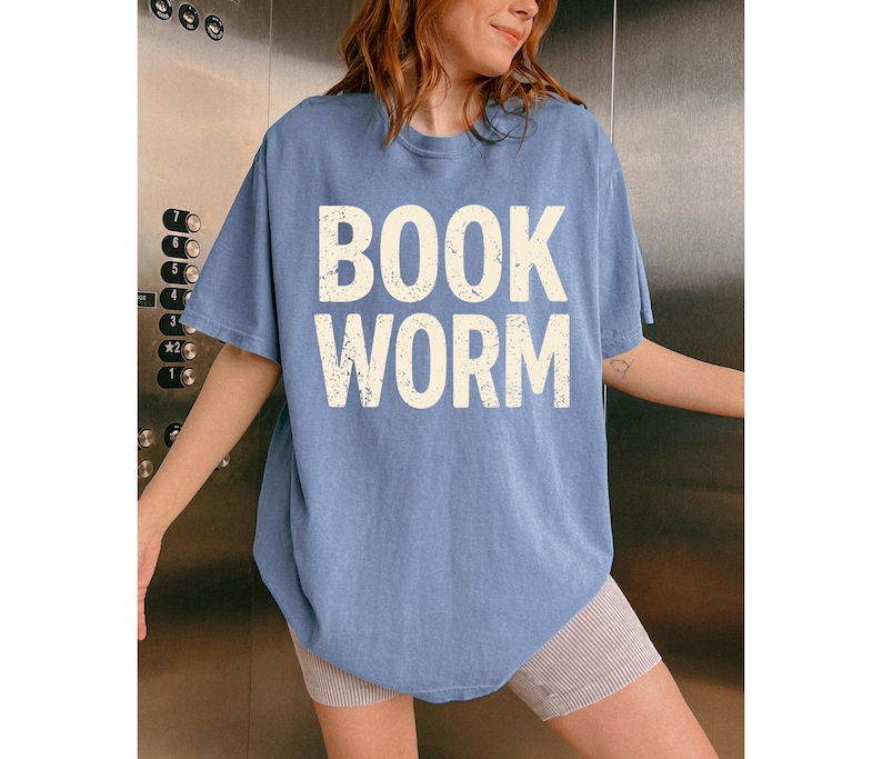 May include: A light blue t-shirt with the words "BOOK WORM" printed in a distressed, off-white font. The shirt is a casual style, with short sleeves and a relaxed fit. The person is wearing striped shorts.