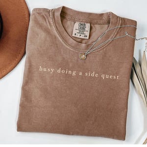May include: A brown Comfort Colors t-shirt with the phrase "busy doing a side quest" printed in white. A silver necklace with a pendant is on top of the shirt. A brown hat is visible in the upper left corner.
