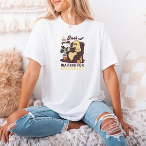 May include: White t-shirt with a cartoon ghost reading a book in an armchair. The graphic includes the text "Just Waiting for Halloween" in a vintage style. The design has a Halloween theme.