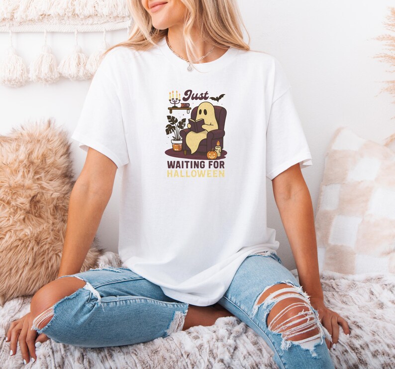 May include: White t-shirt with a cartoon ghost reading a book in an armchair. The graphic includes the text "Just Waiting for Halloween" in a vintage style. The design has a Halloween theme.