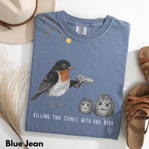 Puede incluir: Una camiseta color azul jean con un gr&aacute;fico de un p&aacute;jaro sosteniendo una pistola apuntando a dos piedras con ojos en forma de X. El texto debajo del gr&aacute;fico dice: "KILLING TWO STONES WITH ONE BIRD." La camiseta tiene cuello redondo.