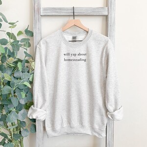 K&ouml;nnte beinhalten: Hellgraues Sweatshirt mit dem schwarzen Text "will yap about homesteading". Das lang&auml;rmlige Rundhals-Shirt h&auml;ngt an einem Holzkleiderb&uuml;gel vor einem wei&szlig;en Hintergrund.
