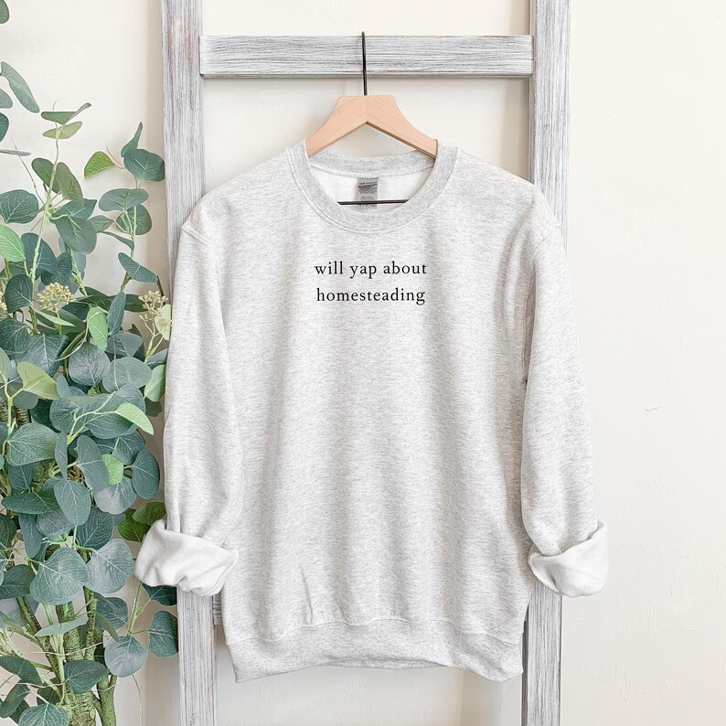 K&ouml;nnte beinhalten: Hellgraues Sweatshirt mit dem schwarzen Text "will yap about homesteading". Das lang&auml;rmlige Rundhals-Shirt h&auml;ngt an einem Holzkleiderb&uuml;gel vor einem wei&szlig;en Hintergrund.