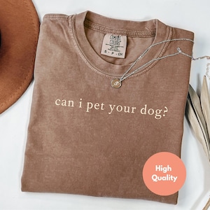 May include: A brown Comfort Colors t-shirt with the phrase "can i pet your dog?" printed in white. The shirt is a crew neck and has a small tag at the neckline. A silver necklace with a pendant is draped across the shirt.