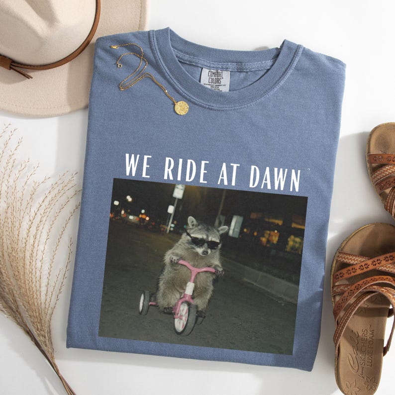 Puede incluir: Una camiseta azul Comfort Colors con el texto "WE RIDE AT DAWN" sobre una foto de un mapache con gafas de sol montando un triciclo rosa. Una cadena de oro, un sombrero y sandalias tambi&eacute;n est&aacute;n en la imagen.