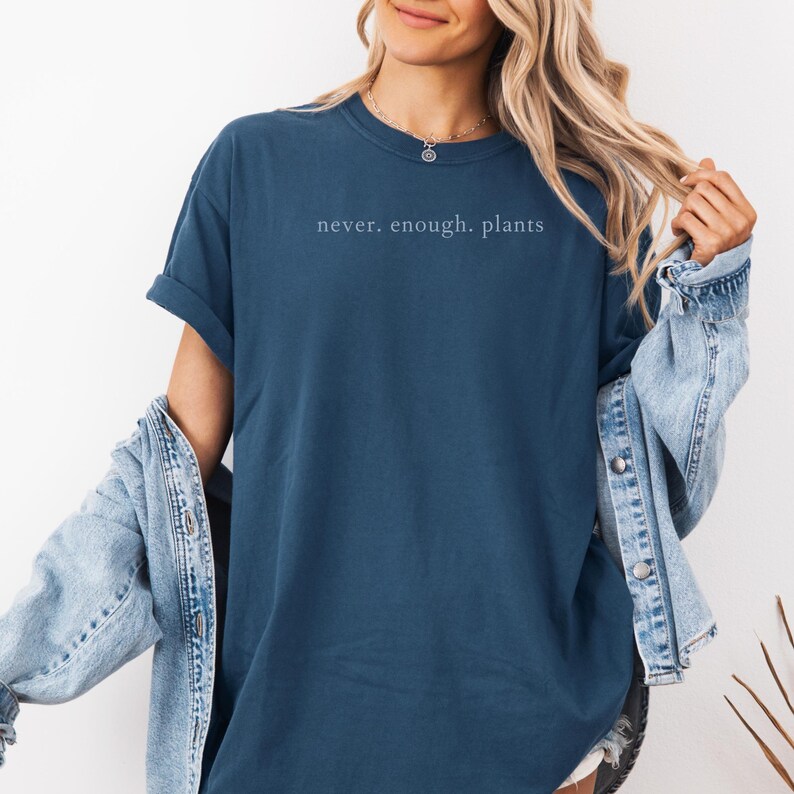 May include: A navy blue t-shirt with the text "never. enough. plants" in a light color. The shirt has short sleeves and a crew neck. A denim jacket is worn over the shirt.