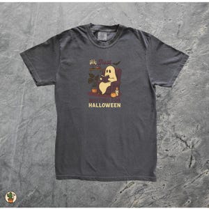 May include: Dark grey t-shirt with a Halloween graphic. The design shows a ghost reading in a chair, with the text "Just Waiting for Halloween". The illustration includes a pumpkin, candles, and a bat.
