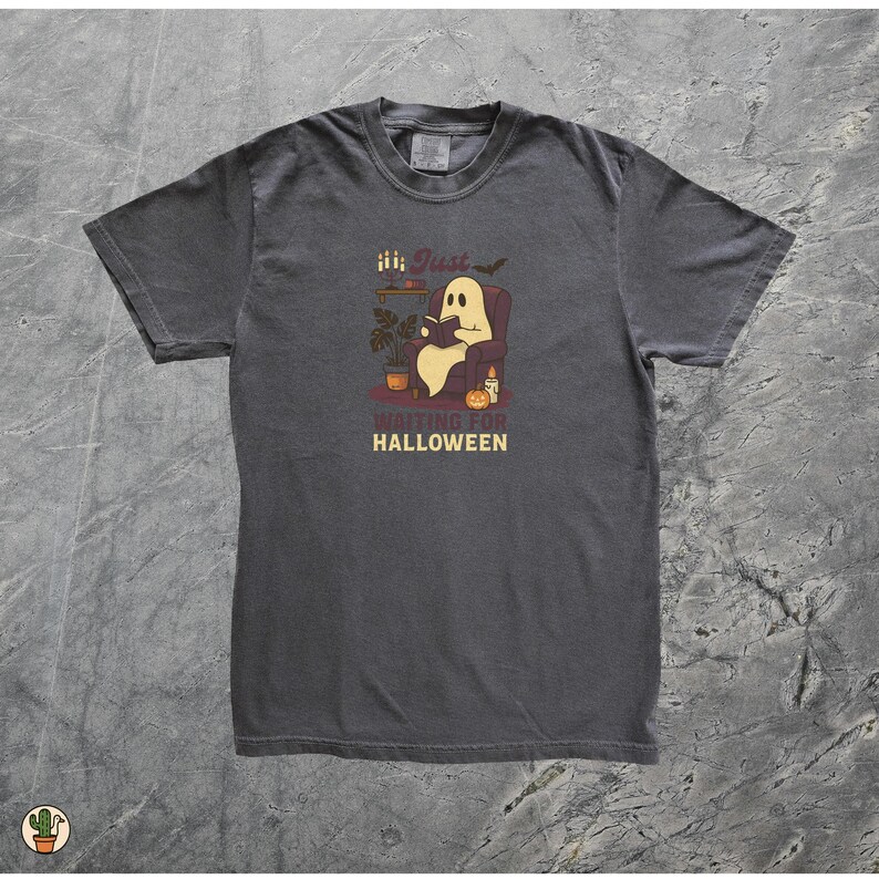 May include: Dark grey t-shirt with a Halloween graphic. The design shows a ghost reading in a chair, with the text "Just Waiting for Halloween". The illustration includes a pumpkin, candles, and a bat.