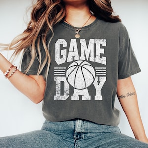 Puede incluir: Camiseta gris oscuro con las palabras "GAME DAY" en letras blancas desgastadas, con un gr&aacute;fico de baloncesto. La camiseta es de estilo casual, adecuada para los fan&aacute;ticos del deporte.