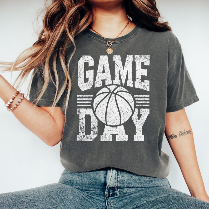 Puede incluir: Camiseta gris oscuro con las palabras "GAME DAY" en letras blancas desgastadas, con un gr&aacute;fico de baloncesto. La camiseta es de estilo casual, adecuada para los fan&aacute;ticos del deporte.