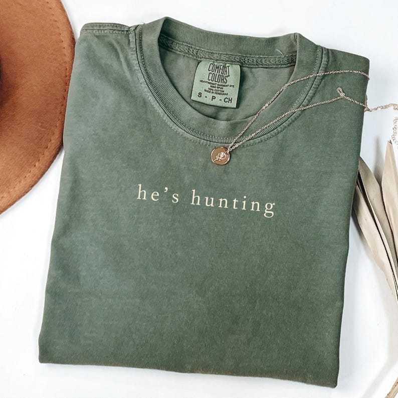 May include: An olive green t-shirt with the phrase "he's hunting" printed in white. A silver necklace with a circular pendant rests on the shirt. The shirt has a Comfort Colors tag.