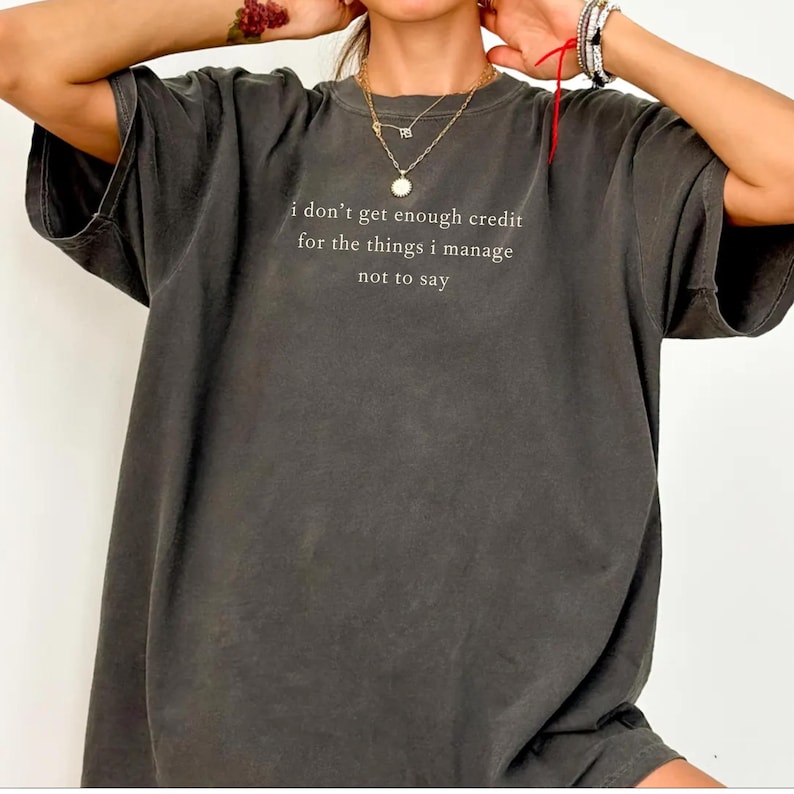 May include: Dark gray t-shirt with the quote "I don't get enough credit for the things I manage not to say" in white lettering. The shirt has a relaxed fit and short sleeves. The overall style is casual and comfortable.