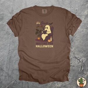 May include: A brown t-shirt featuring a cartoon ghost reading a book in an armchair. The design includes a pumpkin, candles, and the text "Just Waiting for Halloween". A fun Halloween-themed garment.