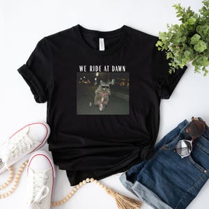 Puede incluir: Camiseta negra con un gr&aacute;fico de un mapache montando un triciclo rosa por la noche, con el texto "WE RIDE AT DAWN". La camiseta se combina con zapatillas blancas, vaqueros azules y gafas de sol.