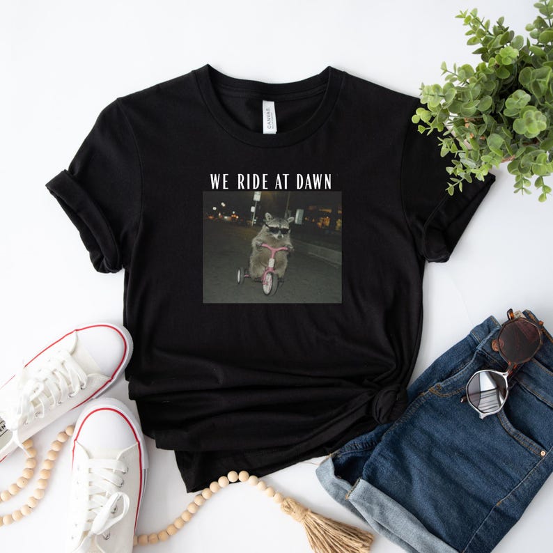 Puede incluir: Camiseta negra con un gr&aacute;fico de un mapache montando un triciclo rosa por la noche, con el texto "WE RIDE AT DAWN". La camiseta se combina con zapatillas blancas, vaqueros azules y gafas de sol.