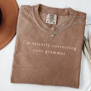 Può includere: Una maglietta Comfort Colors marrone con la frase "i'm silently correcting your grammar" stampata in bianco. Una collana d'argento con un piccolo ciondolo è appoggiata sulla maglietta. La maglietta è piegata ordinatamente su una superficie bianca.