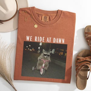 Puede incluir: Camiseta color &oacute;xido con la frase "WE RIDE AT DAWN" sobre una imagen de un mapache con gafas de sol en un triciclo rosa. La camiseta es de la marca Comfort Colors.
