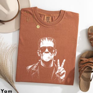 Puede incluir: Una camiseta de manga corta color &oacute;xido con un estampado gr&aacute;fico blanco de un personaje con pernos en la cabeza, usando gafas de sol y soplando una burbuja, mientras hace el signo de la paz. La camiseta tiene cuello redondo.