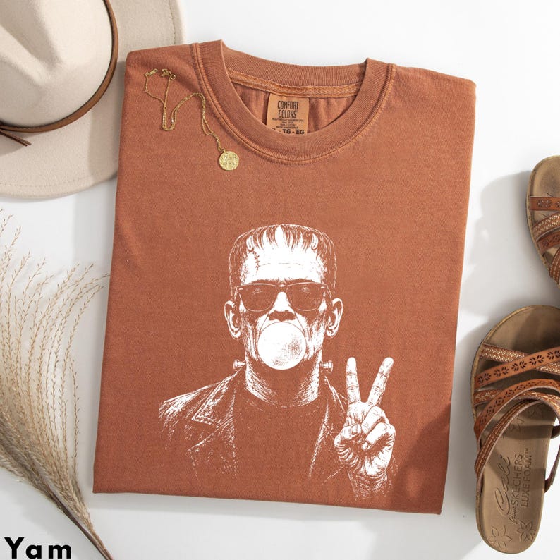 Puede incluir: Una camiseta de manga corta color &oacute;xido con un estampado gr&aacute;fico blanco de un personaje con pernos en la cabeza, usando gafas de sol y soplando una burbuja, mientras hace el signo de la paz. La camiseta tiene cuello redondo.