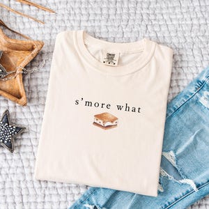 May include: Cream-colored t-shirt with the text "s'more what" and a watercolor illustration of a s'more. The shirt is folded and placed on a textured surface next to a pair of blue jeans.