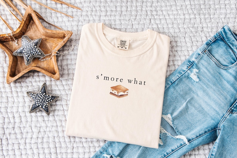 May include: Cream-colored t-shirt with the text "s'more what" and a watercolor illustration of a s'more. The shirt is folded and placed on a textured surface next to a pair of blue jeans.