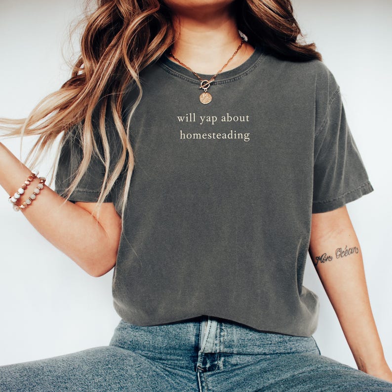 K&ouml;nnte beinhalten: Dunkelgraues T-Shirt mit dem wei&szlig;en Text "will yap about homesteading". Das Shirt hat eine lockere Passform und wird mit einer blauen Jeans getragen. Eine goldene Halskette und ein Armband sind ebenfalls zu sehen.
