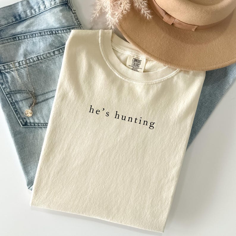 May include: Cream-colored t-shirt with the text "he's hunting" in black, paired with light blue denim jeans, a tan hat, and a silver necklace. The shirt is a Comfort Colors brand.